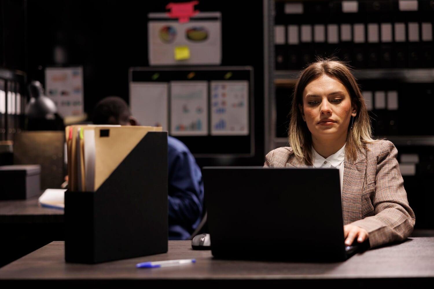Mulher analisando dados em laptop em ambiente corporativo com pastas e gráficos ao fundo